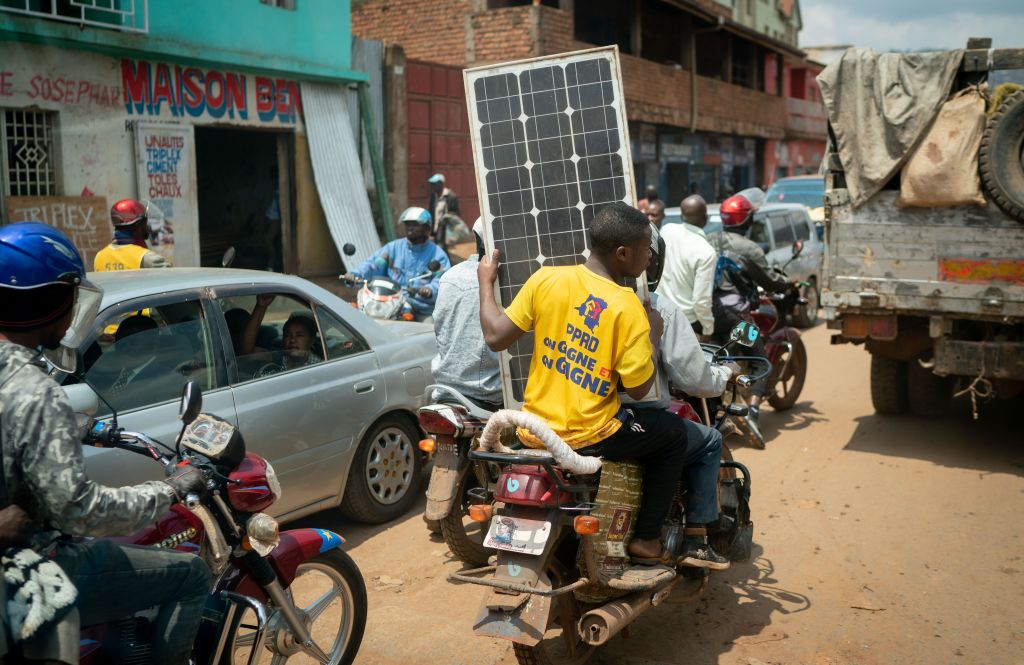 A man holds a solar panel on the back of a motorcycle weaving through dense traffic.
