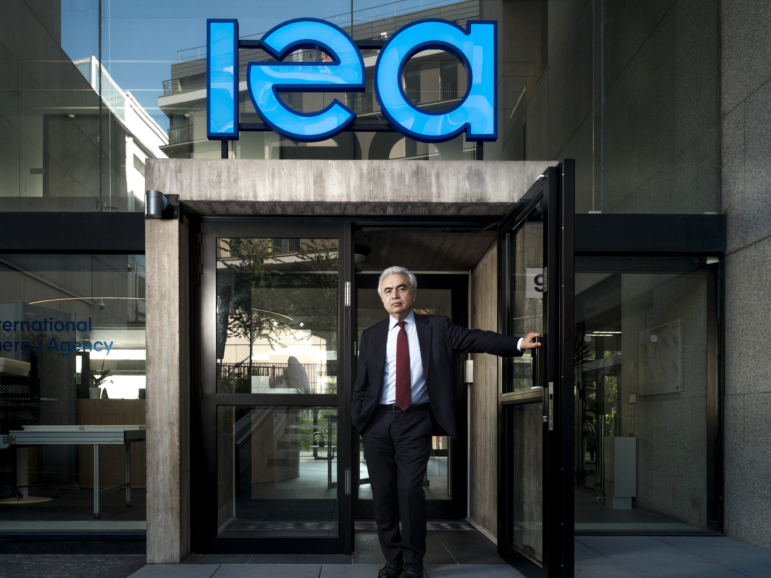 A man stands holding open a glass door, looking at the camera, with a sign that says "IEA" in big blue letters above the door. 