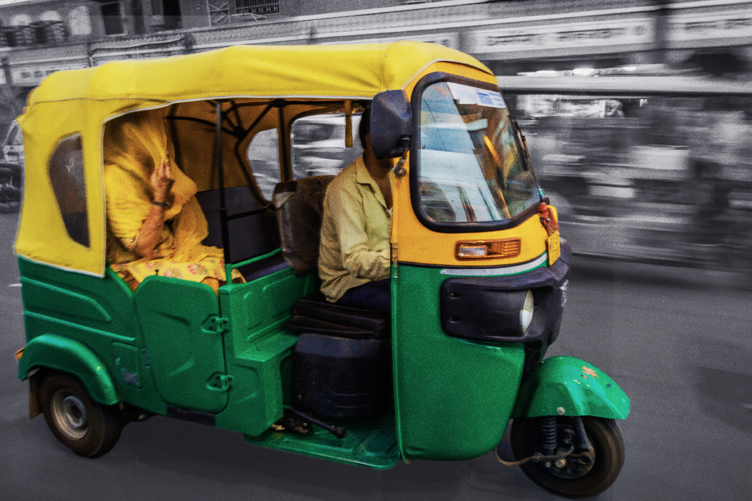 A man drives a three-wheeled auto rickshaw through the streets of Rajasthan, India. The auto rickshaw is yellow and green and the background is black and white. A woman wearing yellow sits in the backseat, her dupatta covering her face.