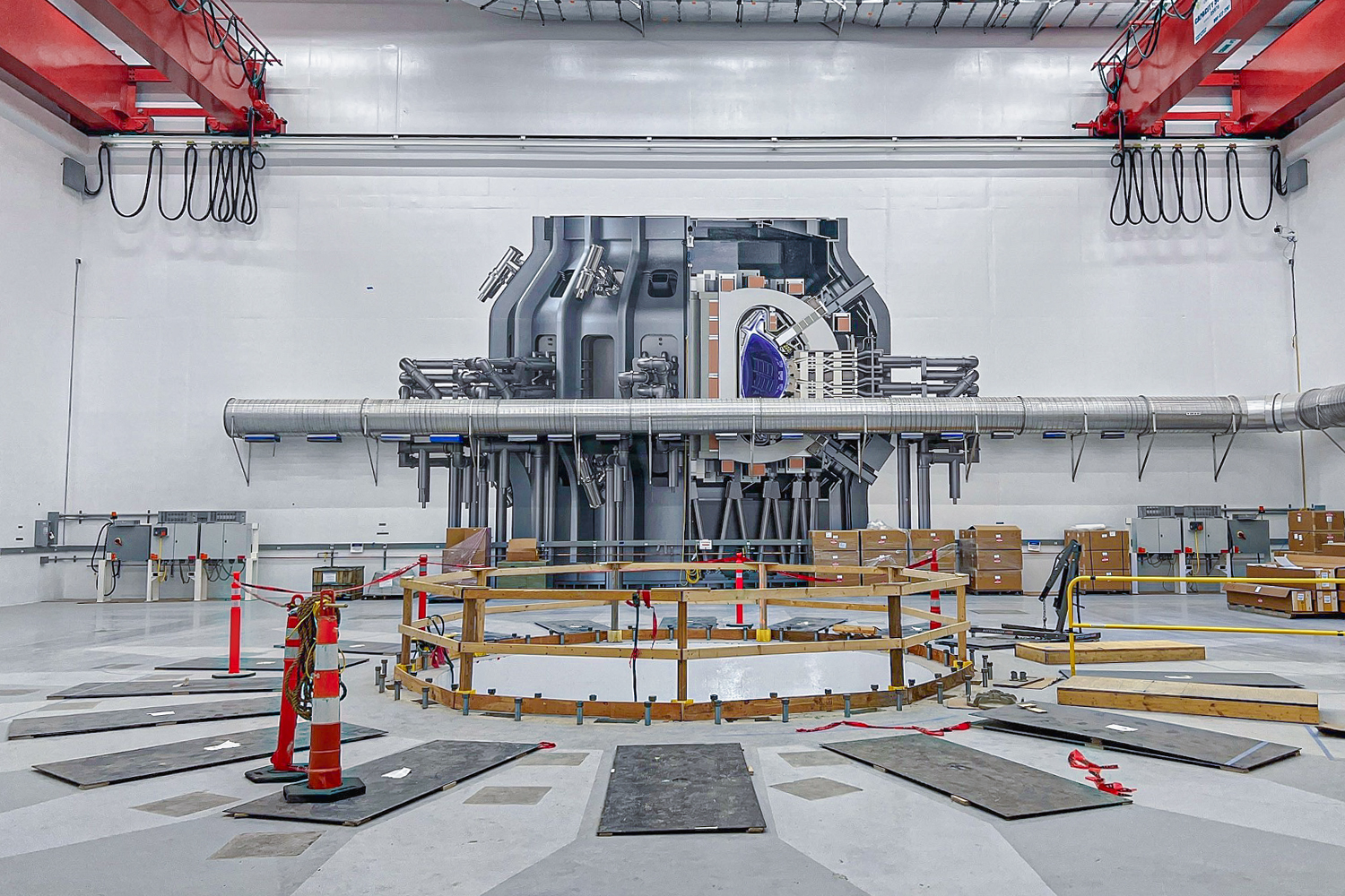 Inside a nuclear fusion facility that is being constructed. There is a large hole in the concrete floor where the reactor will eventually go, and a drawing of the reactor on the wall opposite the hole.