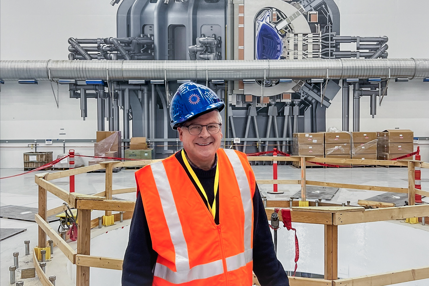 Reporter Bill Spindle in an orange construction vest and hard hat in front of a hole in a concrete floor where a nuclear fusion reactor will eventually go. 