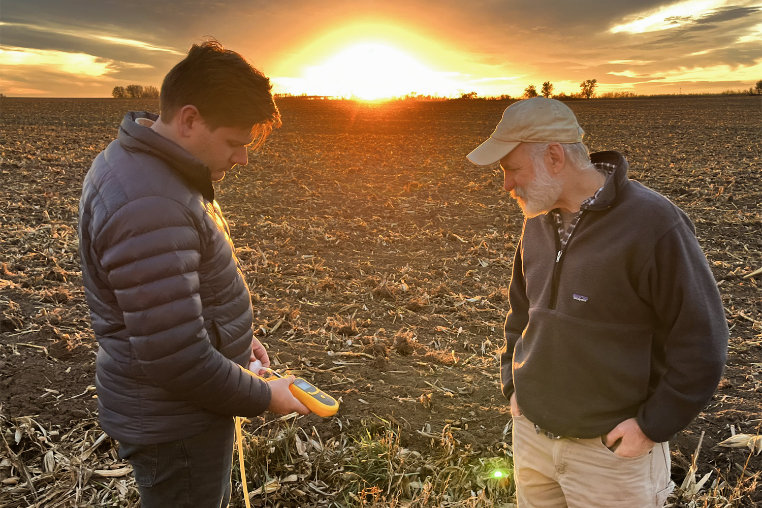 Two men stand in a field, looking at a gas gauge. The sun sets in the background