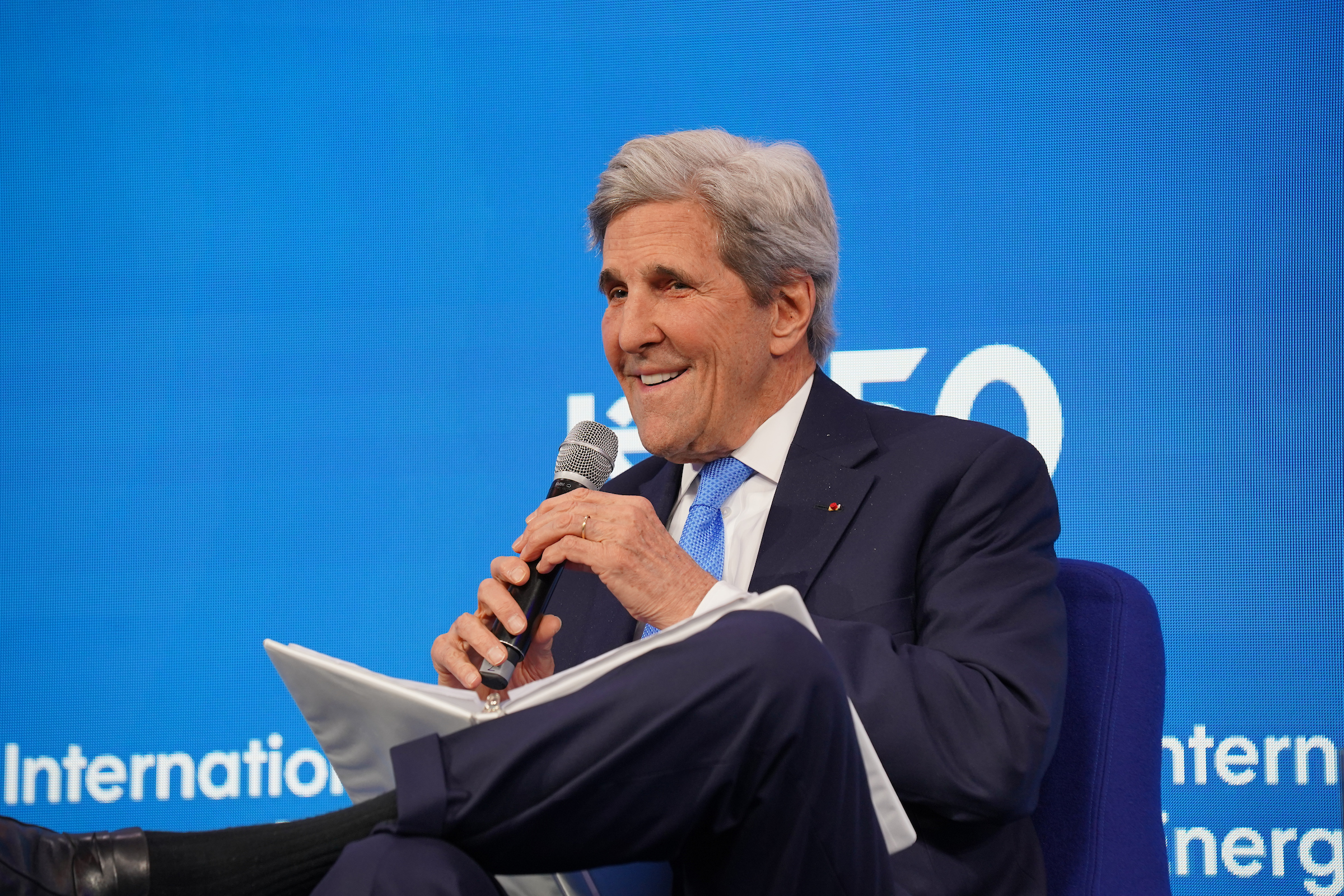 John Kerry speaks into a microphone in front of a blue backdrop. 