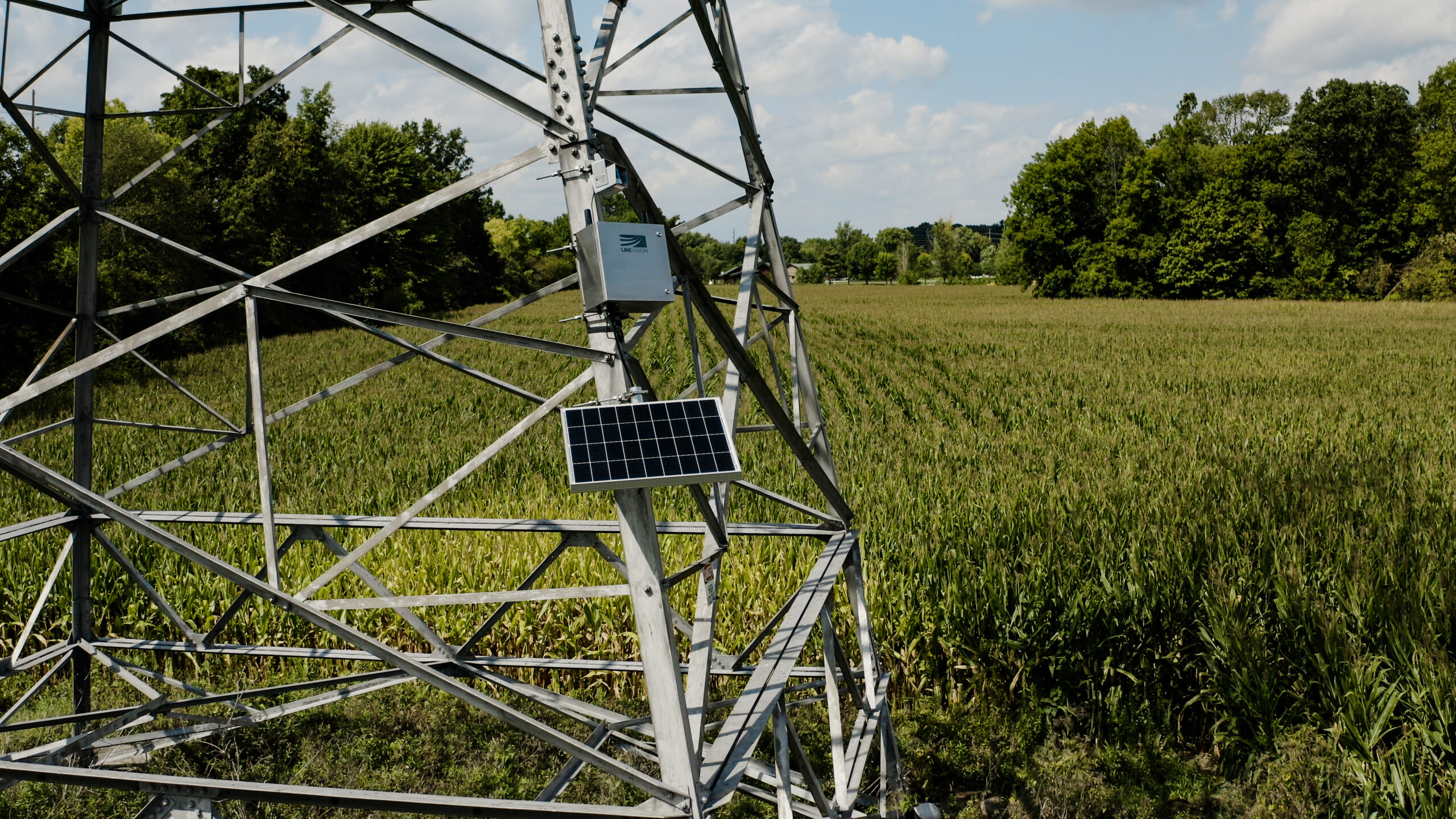 A solar panel powered-sensor sits on a power line in an agricultural field.