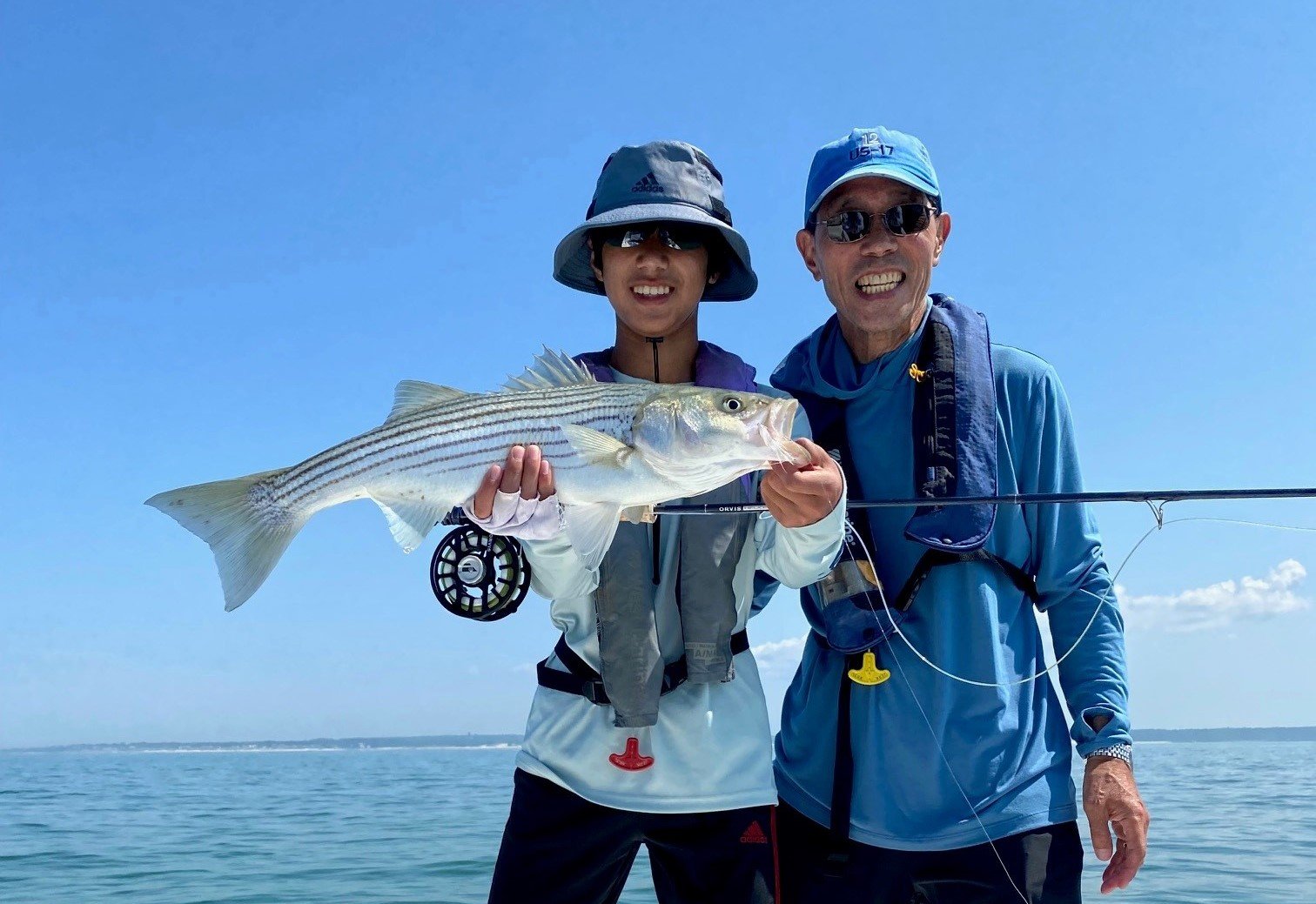 A young man in a bucket hat smiles and holds up a silver striped fish, while an older man in a blue baseball cap smiles next to him. Behind them is blue sky and a large expanse of water.