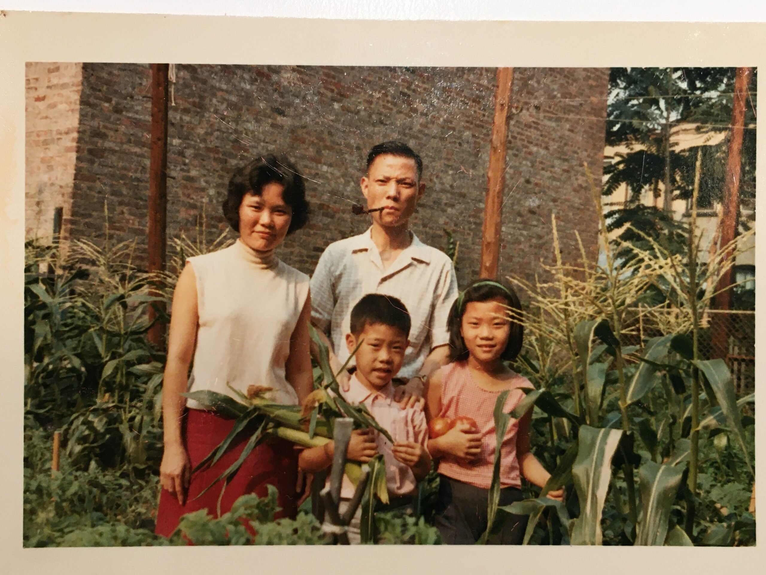An old photograph of a family — a mother, a father and a little boy and a little girl — standing in a small garden in front of a brick building.