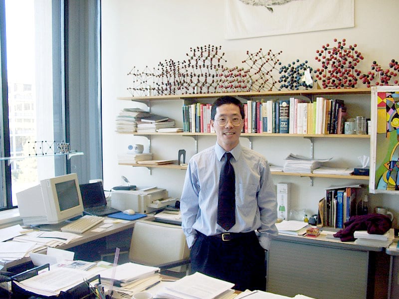 A young man stands with hands in his pockets in an office with stacks of paper everywhere, models of molecules lining a bookshelf, and a clunky early 2000s computer on the desk.