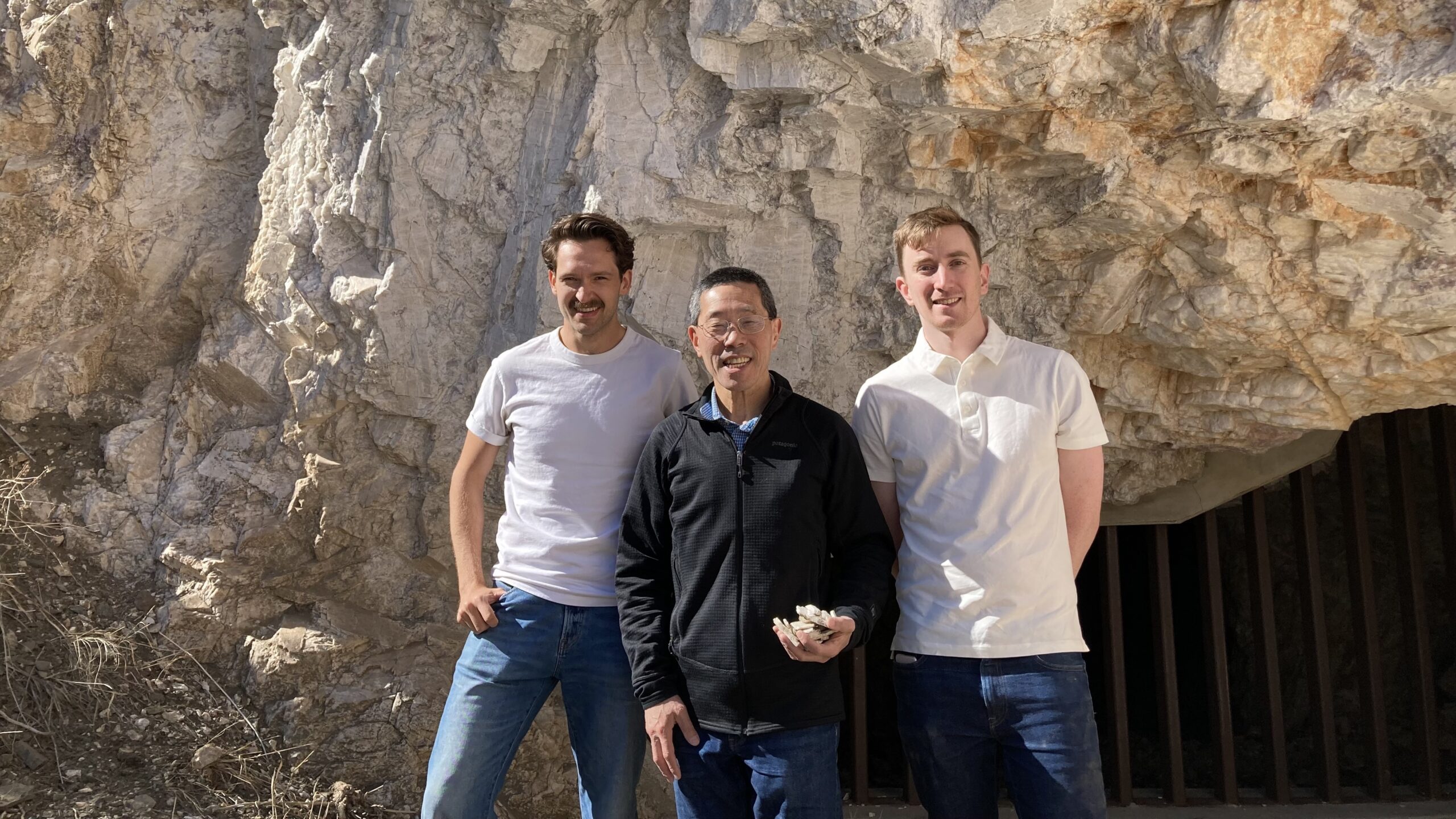 Three men stand in front of a sheer rock face.