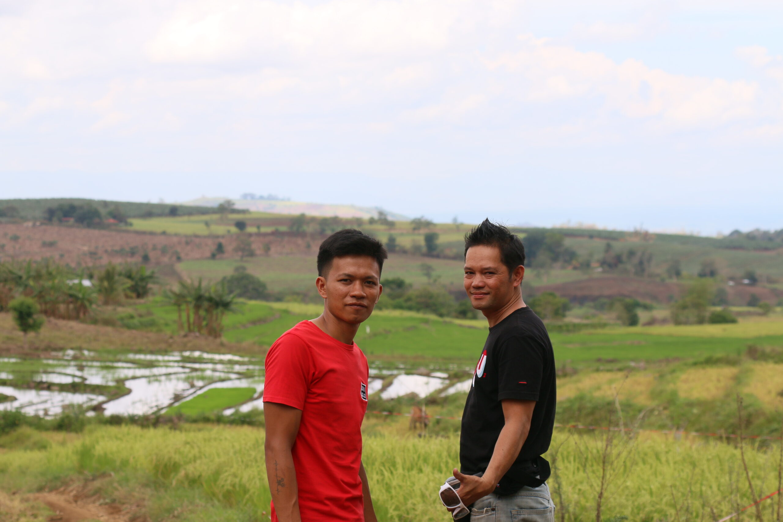 Two men stand in front of a field of green rice paddies behind them looking at the camera in front of them. 