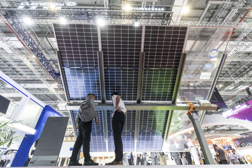 Two men stand under a large solar array in a giant conference center space. Photo shot from floor level, looking up.