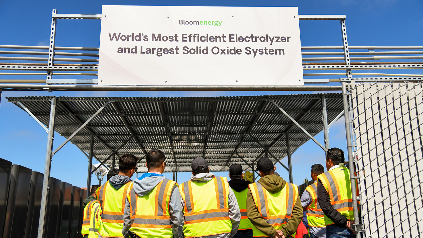 A group of people wearing yellow safety vests gather under a sign that says "Bloom Energy: World's Most Efficient Electrolyzer and Largest Solid Oxide System."