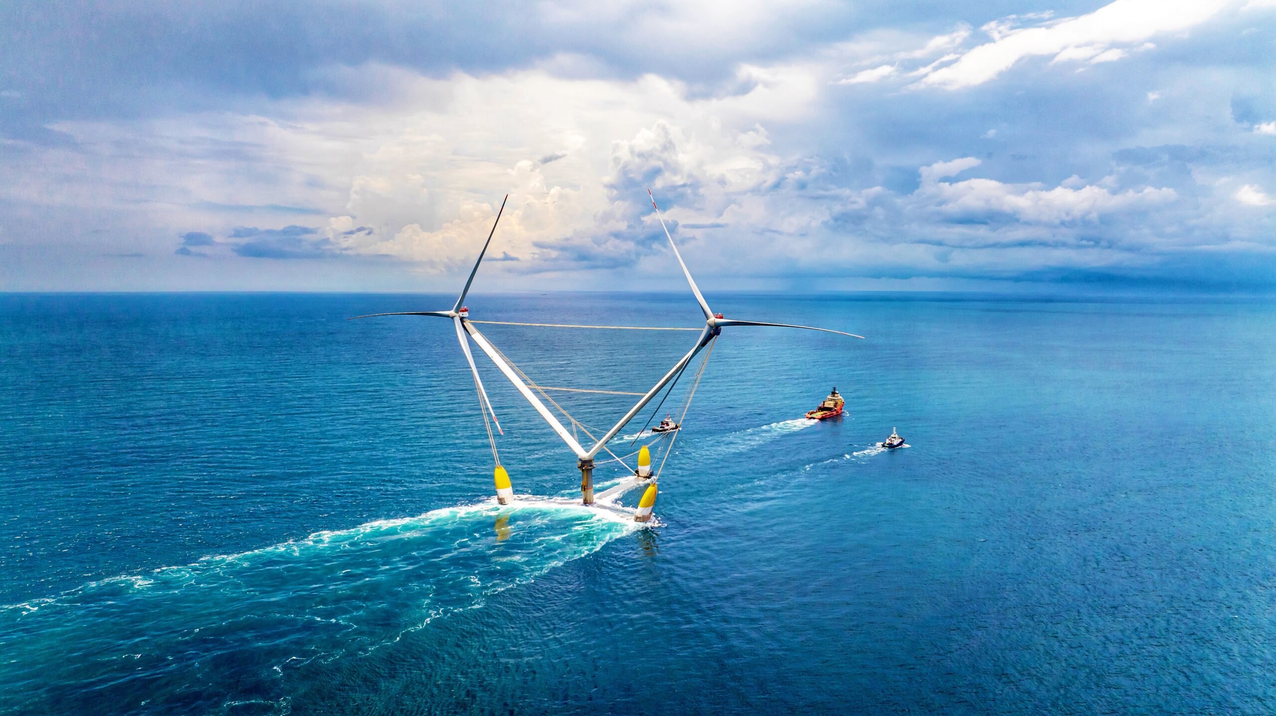 A photograph of a red tug boat pulling a double-headed wind turbine out to sea.